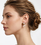 A close-up side profile of a woman with brown hair in an updo, wearing a pair of 18K gold-plated drop earrings with a brown oval tiger's eye gemstone and small white pearl drops. BUCKLE Collective