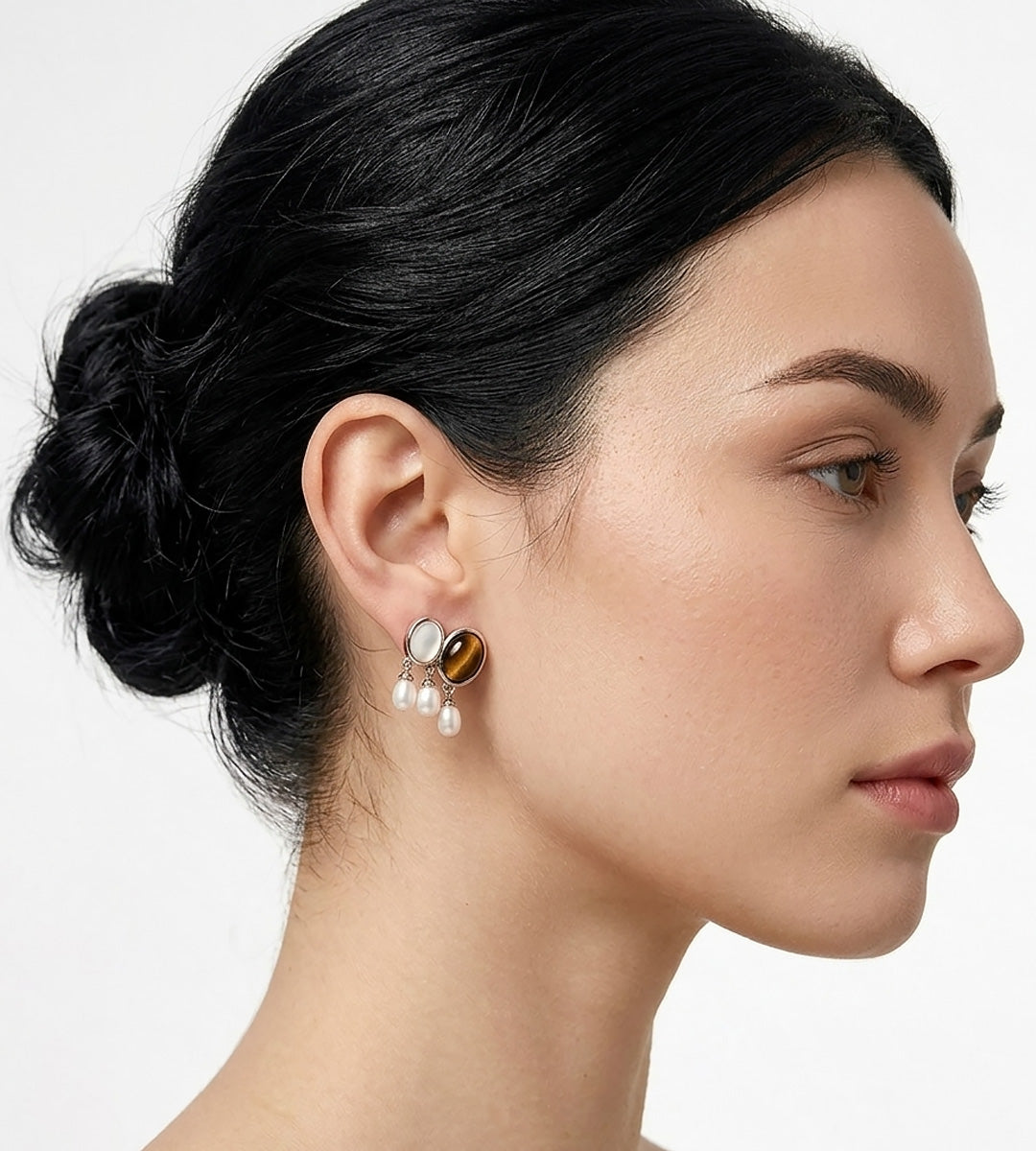 A close-up side profile of a woman with dark hair in a bun, wearing a unique drop earring featuring a round tiger's eye gemstone, mother-of-pearl, and three small natural freshwater white pearls. BUCKLE Collective
