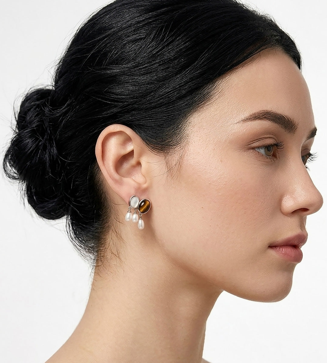 A close-up side profile of a woman with dark hair in a bun, wearing a unique drop earring featuring a round tiger's eye gemstone, mother-of-pearl, and three small natural freshwater white pearls.