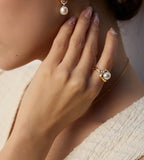 Gold-tone square pearl ring worn on a hand resting beside the neck, styled with a pearl drop earring and cream top.