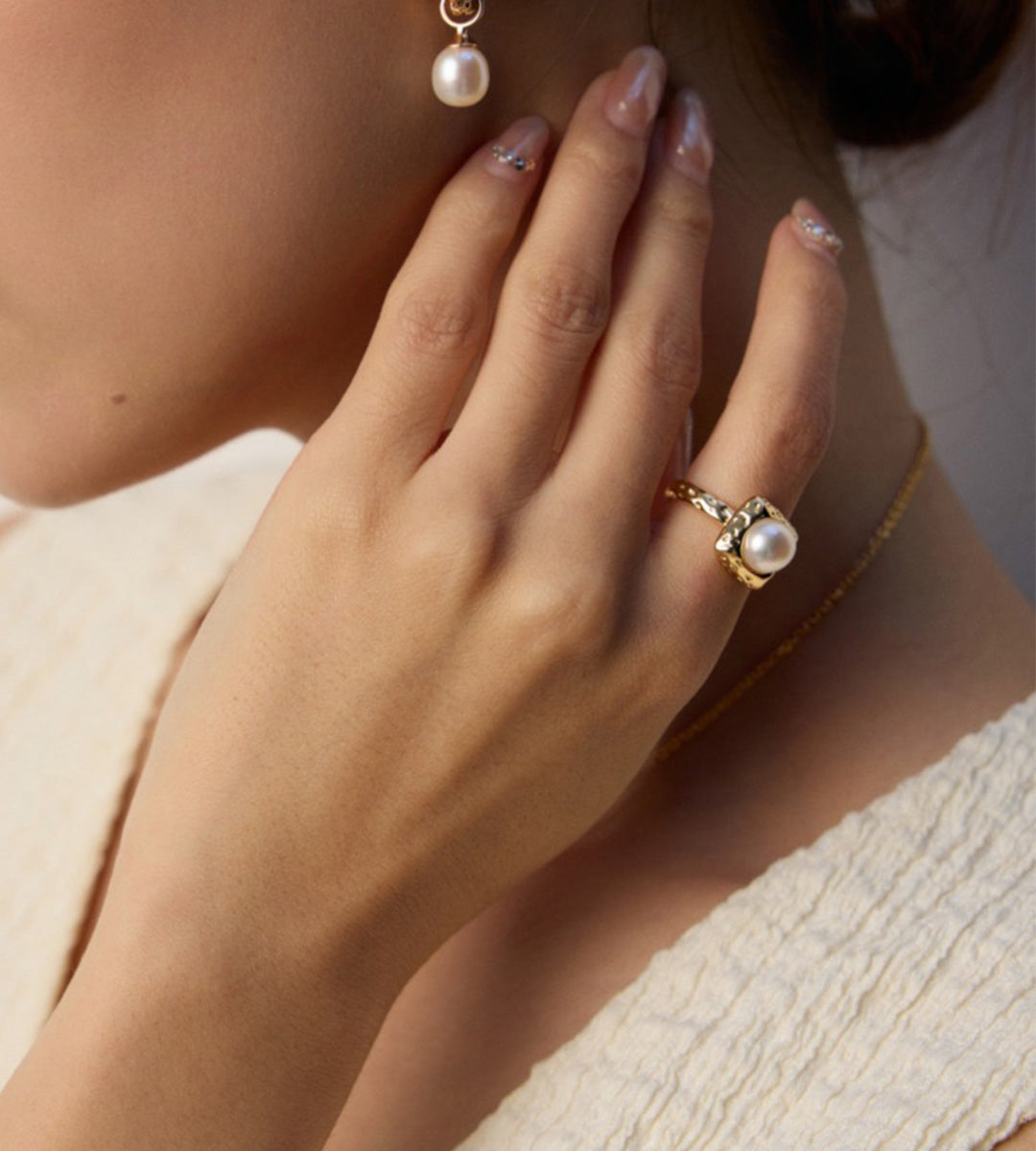 Gold-tone square pearl ring worn on a hand resting beside the neck, styled with a pearl drop earring and cream top.