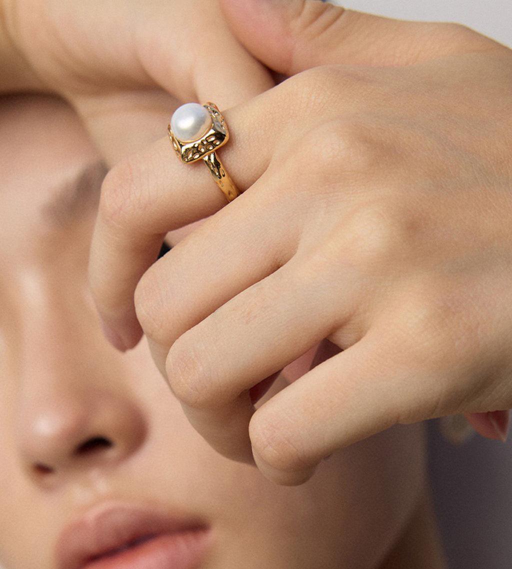 Close-up of a model wearing a gold-tone square pearl ring, showing the raised pearl and hammered-texture frame.