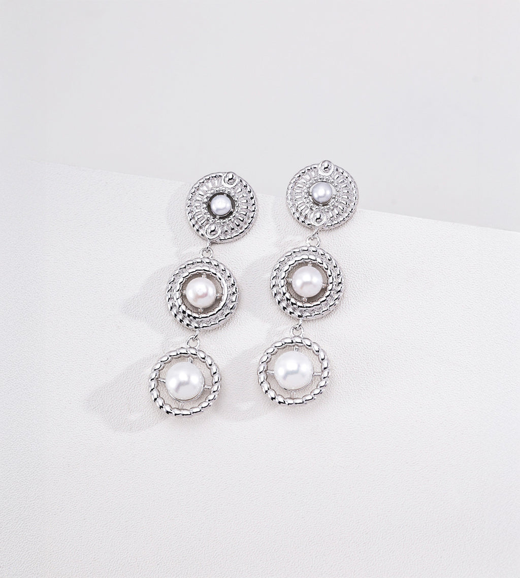 Silver-tone three-tier pearl drop earrings with pearl centres and textured circular frames on a white display.