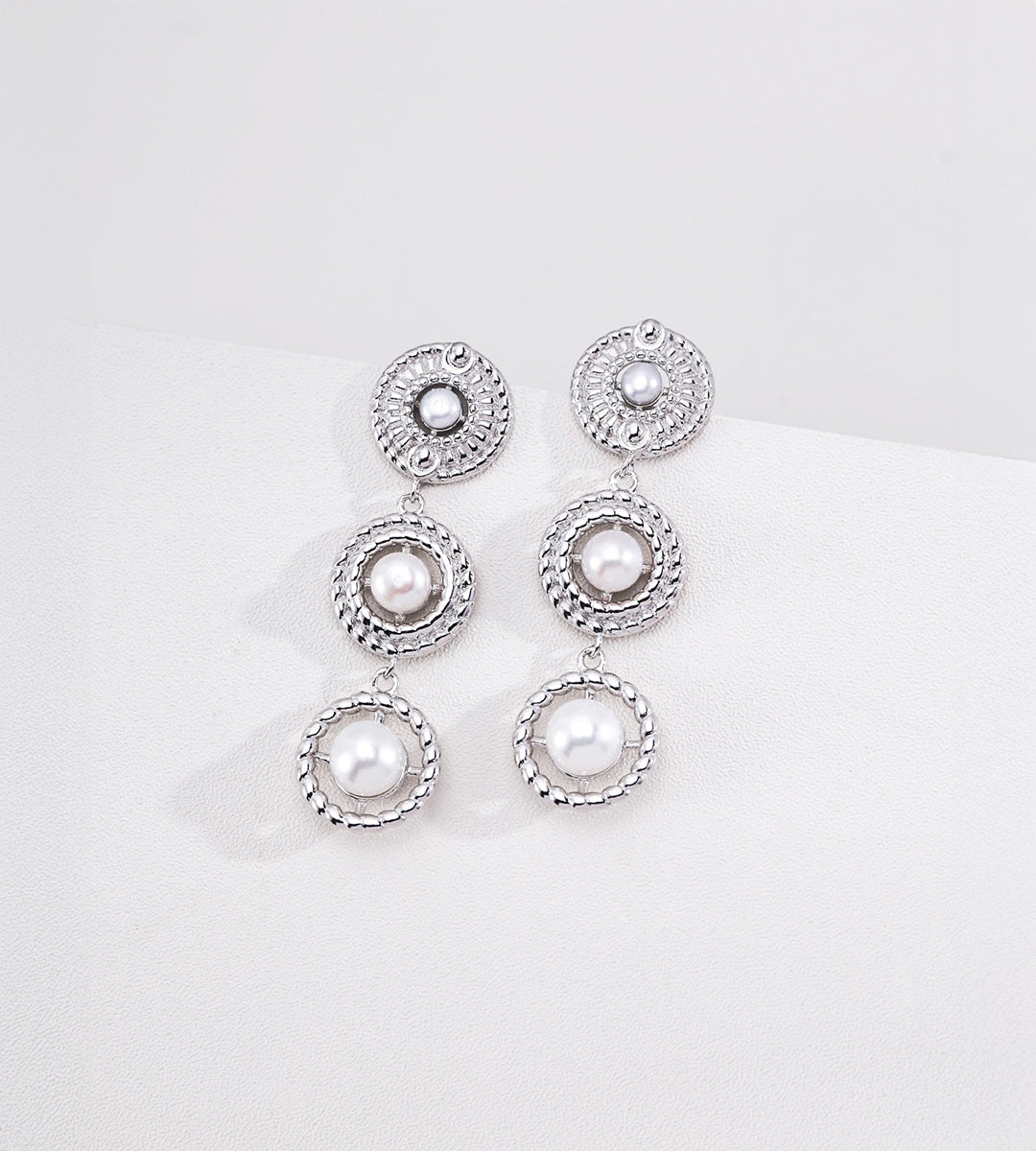 Silver-tone three-tier pearl drop earrings with pearl centres and textured circular frames on a white display.