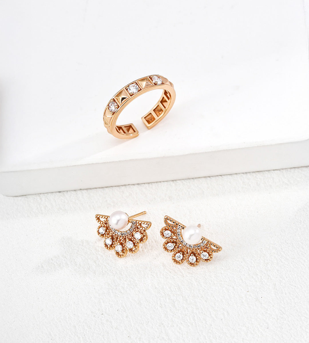 Gold-tone rivet and cubic zirconia open ring displayed above fan-shaped pearl and clear-stone earrings on white blocks.