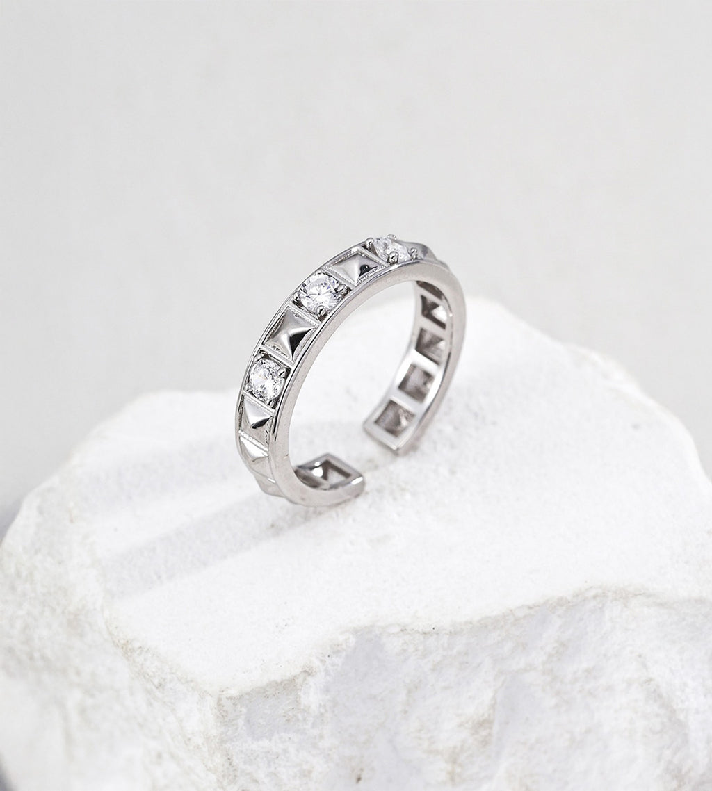Silver-tone open ring with alternating pyramid-shaped rivets and round cubic zirconia, standing on a white stone block.