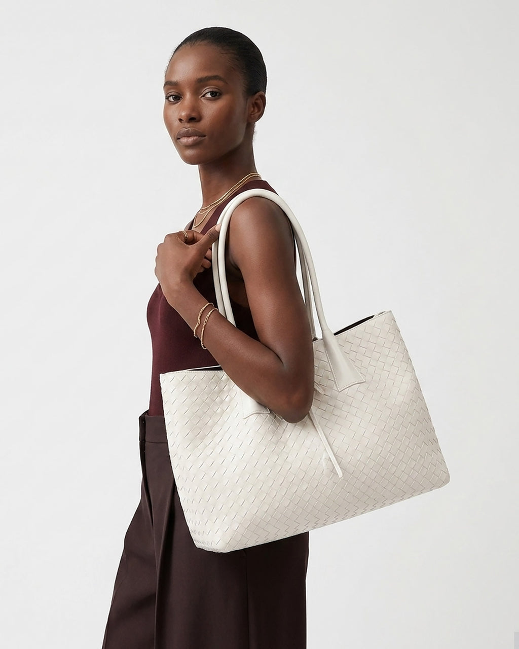 A woman wearing a burgundy sleeveless top and wide dark brown pants carries a large off-white woven tote bag with white shoulder straps