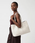 A woman wearing a burgundy sleeveless top and wide dark brown pants carries a large off-white woven tote bag with white shoulder straps