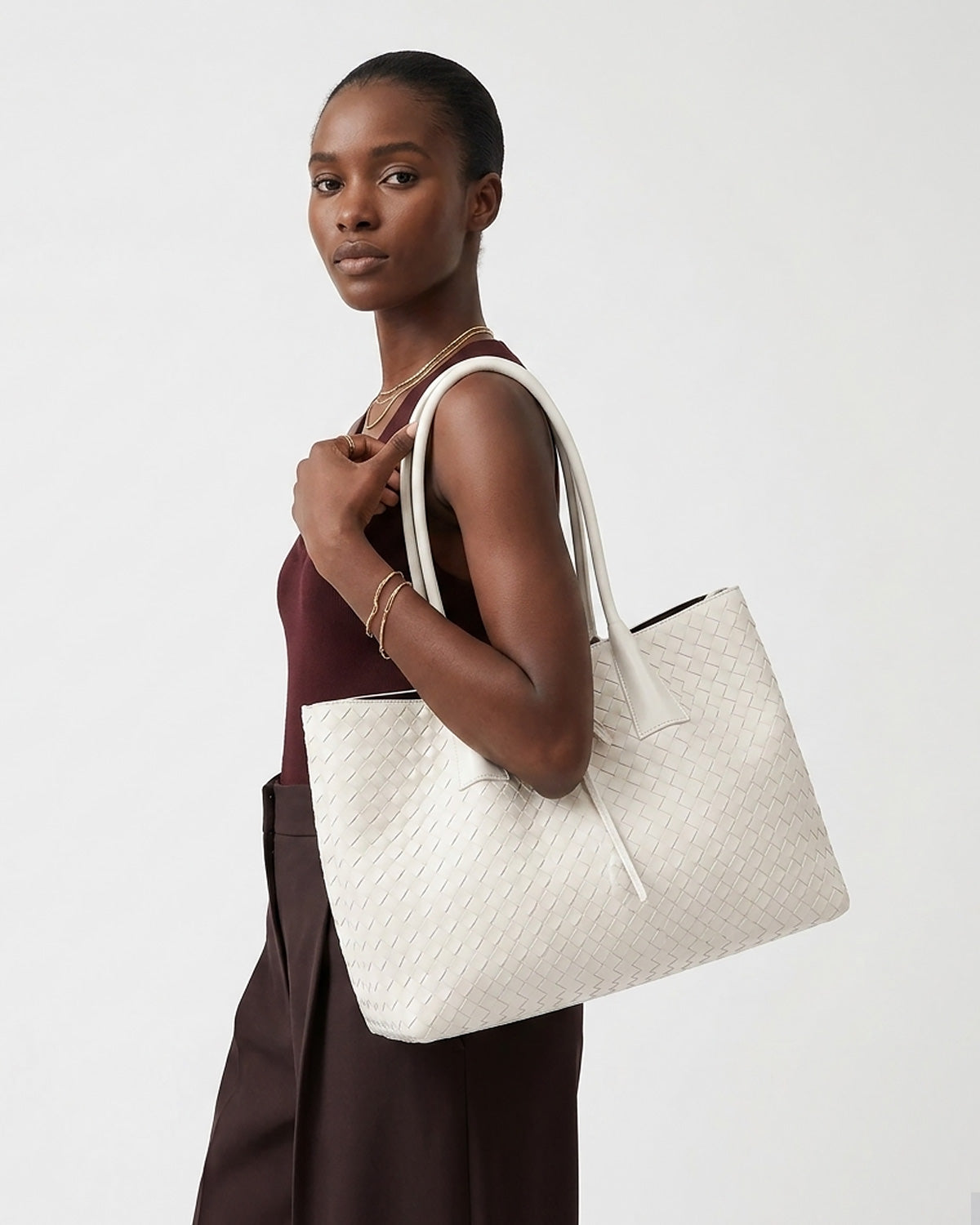 A woman wearing a burgundy sleeveless top and wide dark brown pants carries a large off-white woven tote bag with white shoulder straps
