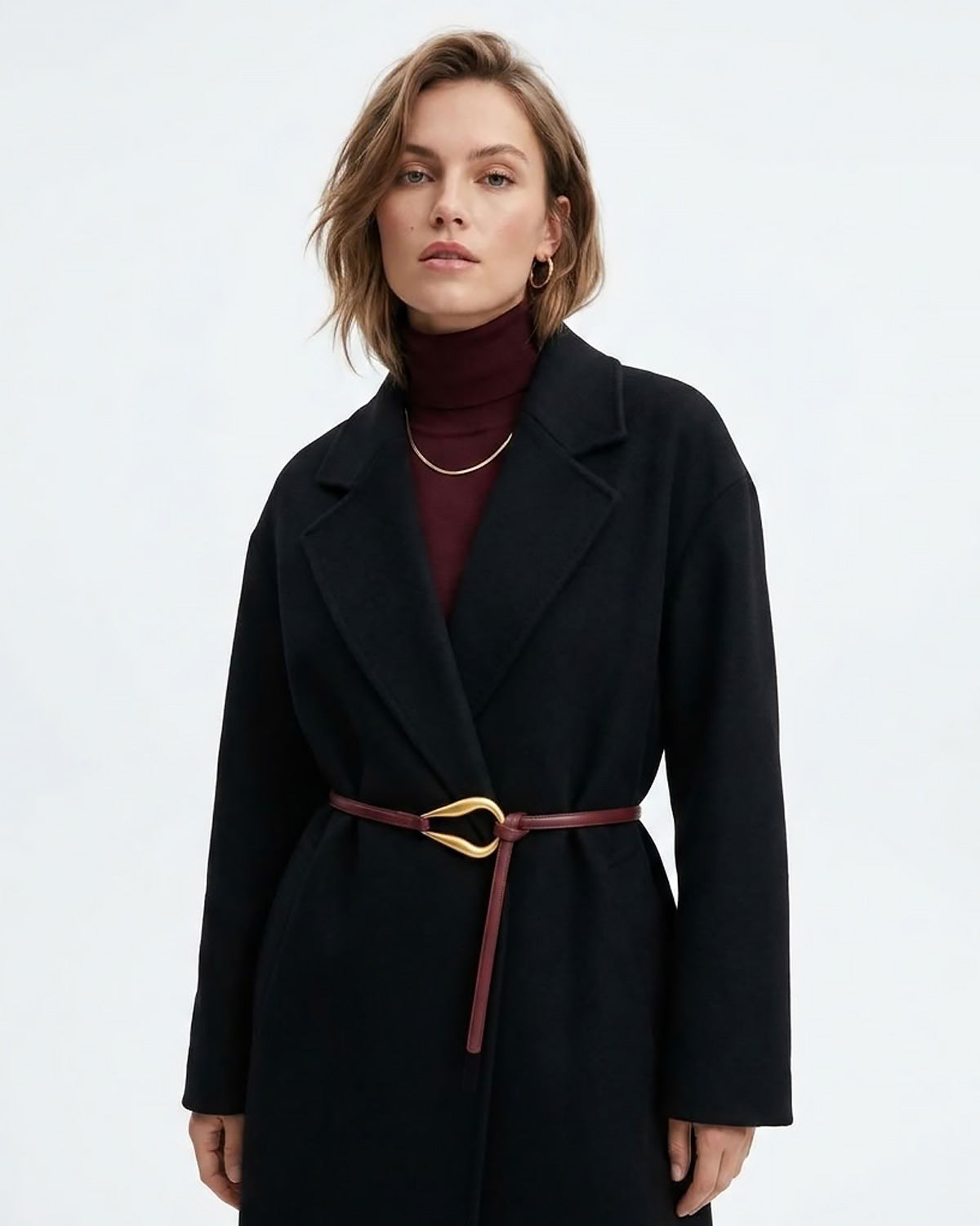 A woman wearing a black belted coat over a burgundy turtleneck sweater, styled with a Slim Knotted Burgundy Leather Belt with Gold Loop