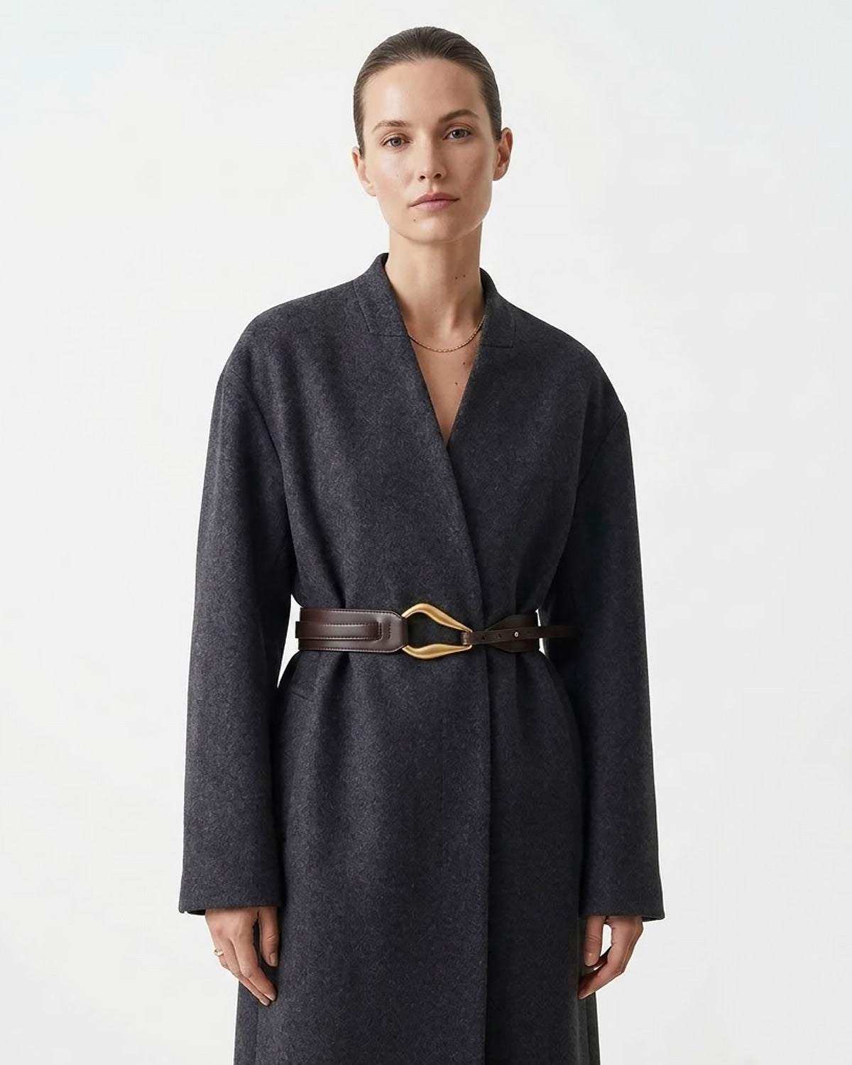 A woman in a dark charcoal gray wrap coat with a Coffee-colored Leather Belt with a sculptural gold-toned hook clasp by BUCKLE Collective