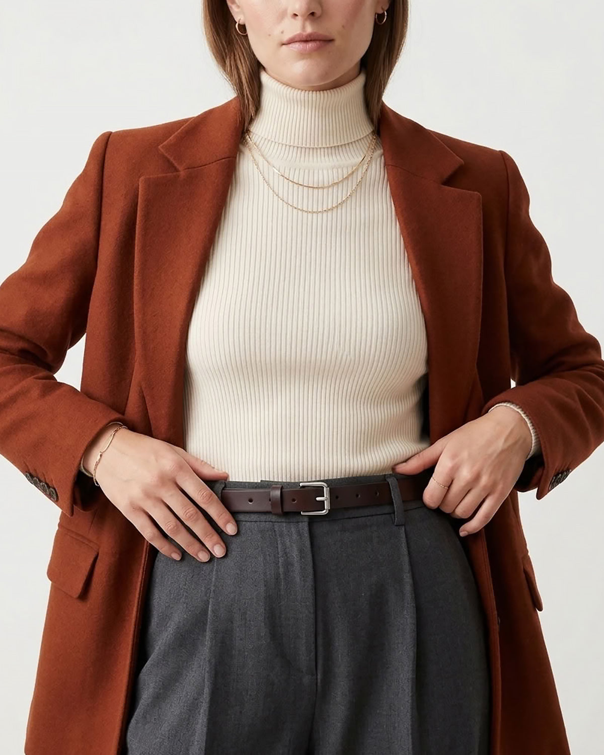 A woman wearing a rust-brown tailored blazer over a cream ribbed turtleneck, paired with dark gray trousers and Leather Belt with a rectangular buckle