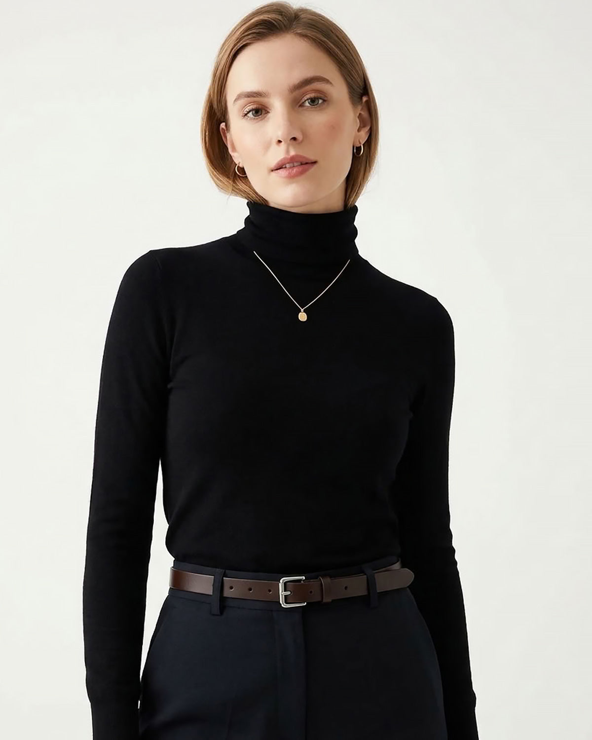 A woman wearing a black ribbed turtleneck sweater, dark trousers, and a brown leather belt
