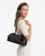 A woman wearing a cream-colored knit top and trousers carries a black leather shoulder bag with a long strap and a decorative black pom-pom detail