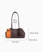 A dark brown leather handbag shown on a light gray background with overlaid dimension lines and measurements for width, height, and handle length. A small orange phone is shown next to the bag for scale.