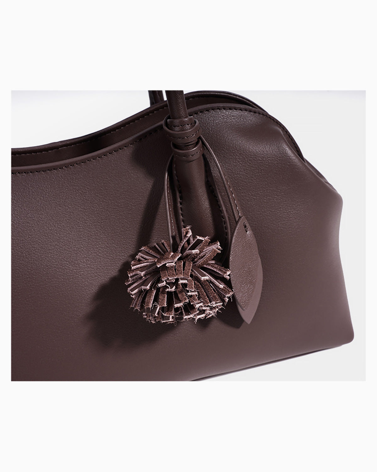 A close-up of a coffee-colored leather handbag featuring a decorative matching leather tassel charm and small leather tags