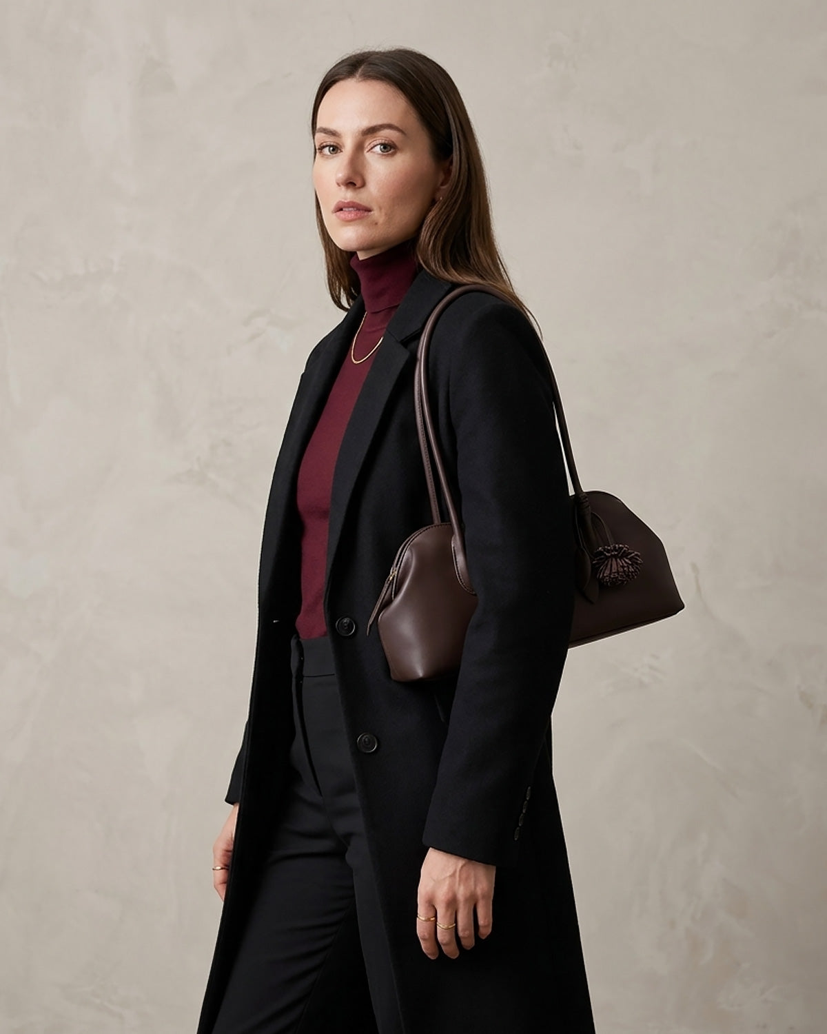 A woman wearing a long black coat over a burgundy turtleneck, carrying a dark brown leather shoulder bag against a textured light gray wall.