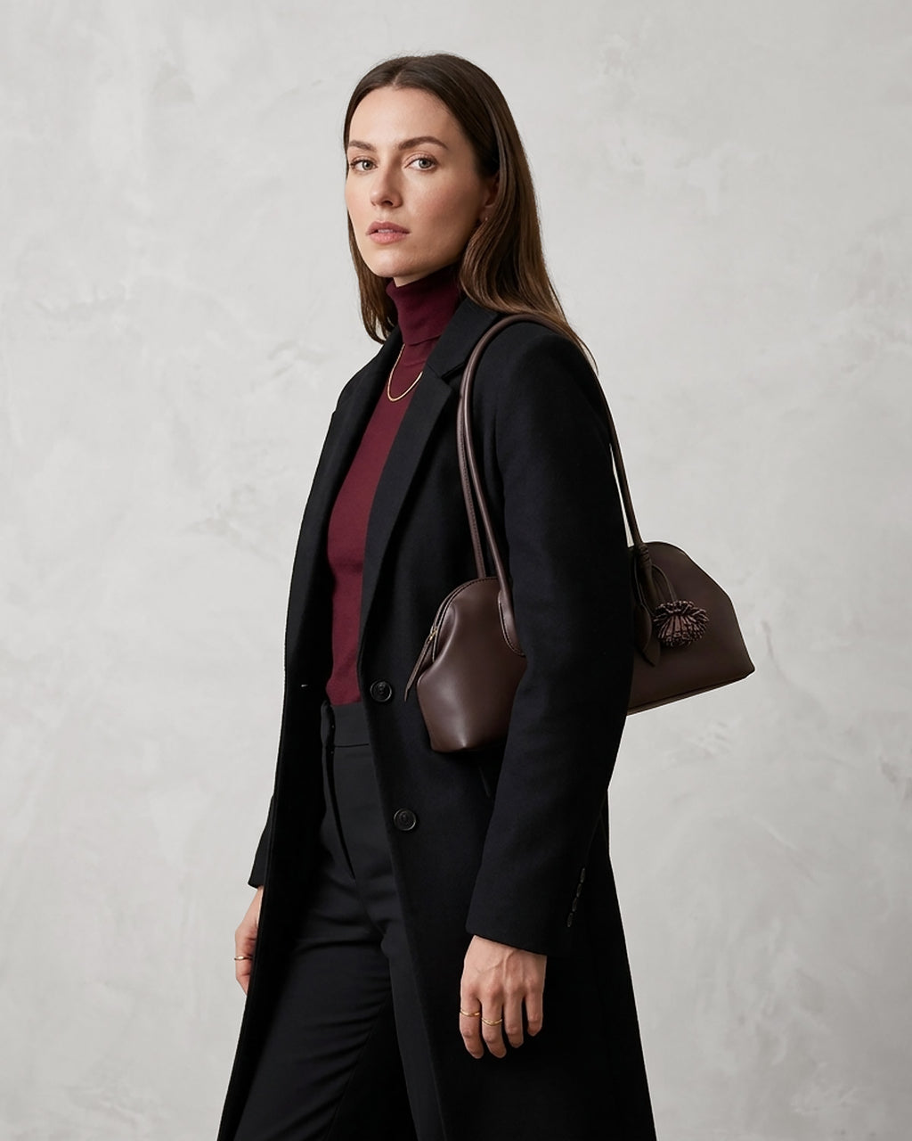 A woman wearing a long black coat over a burgundy turtleneck, carrying a dark brown leather shoulder bag against a textured light gray wall.