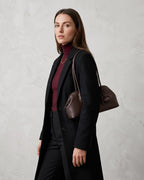 A woman wearing a long black coat over a burgundy turtleneck, carrying a dark brown leather shoulder bag against a textured light gray wall.