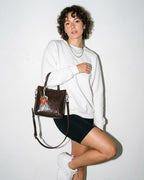 A woman in a chic casual outfit carries a Coffee Brown Genuine Leather Mini Tote Crossbody Bag with Strawberry and Floral Charm