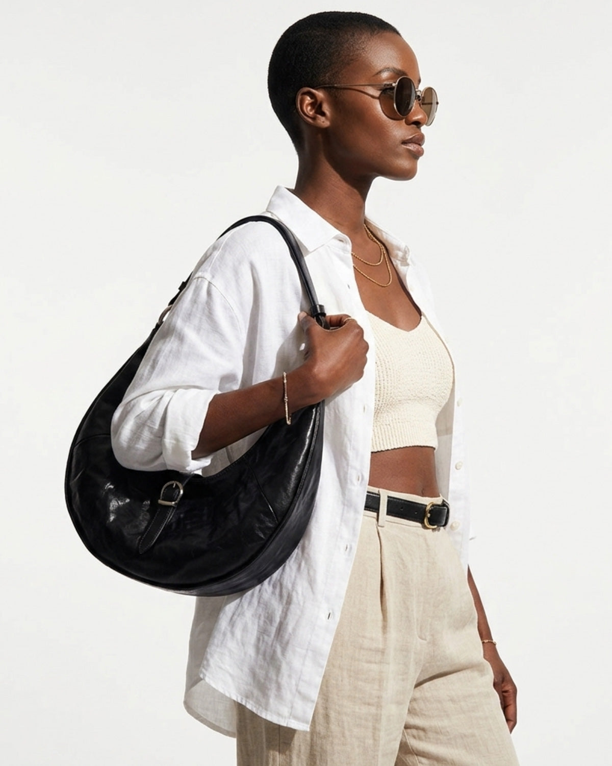 A stylish woman in sunglasses and a relaxed summer outfit carries a Black Leather Dumpling Bag with Adjustable Strap & Buckle Accent