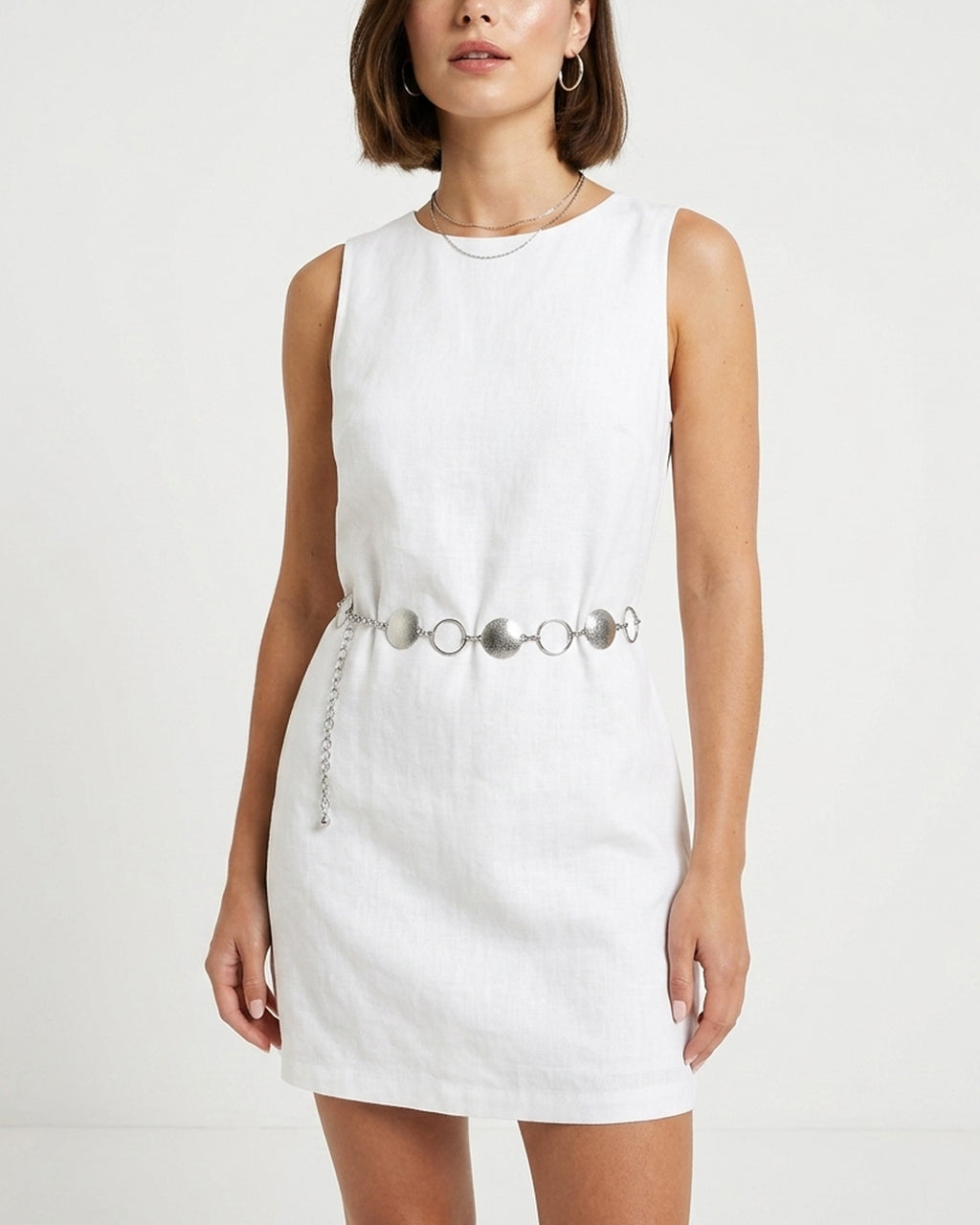 A woman wearing a sleeveless white linen dress with a silver chain belt featuring circular metal accents | BUCKLE Collective