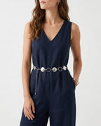 A woman wearing a navy blue sleeveless jumpsuit with a V-neckline and a decorative silver chain belt with rings and discs | BUCKLE Collective