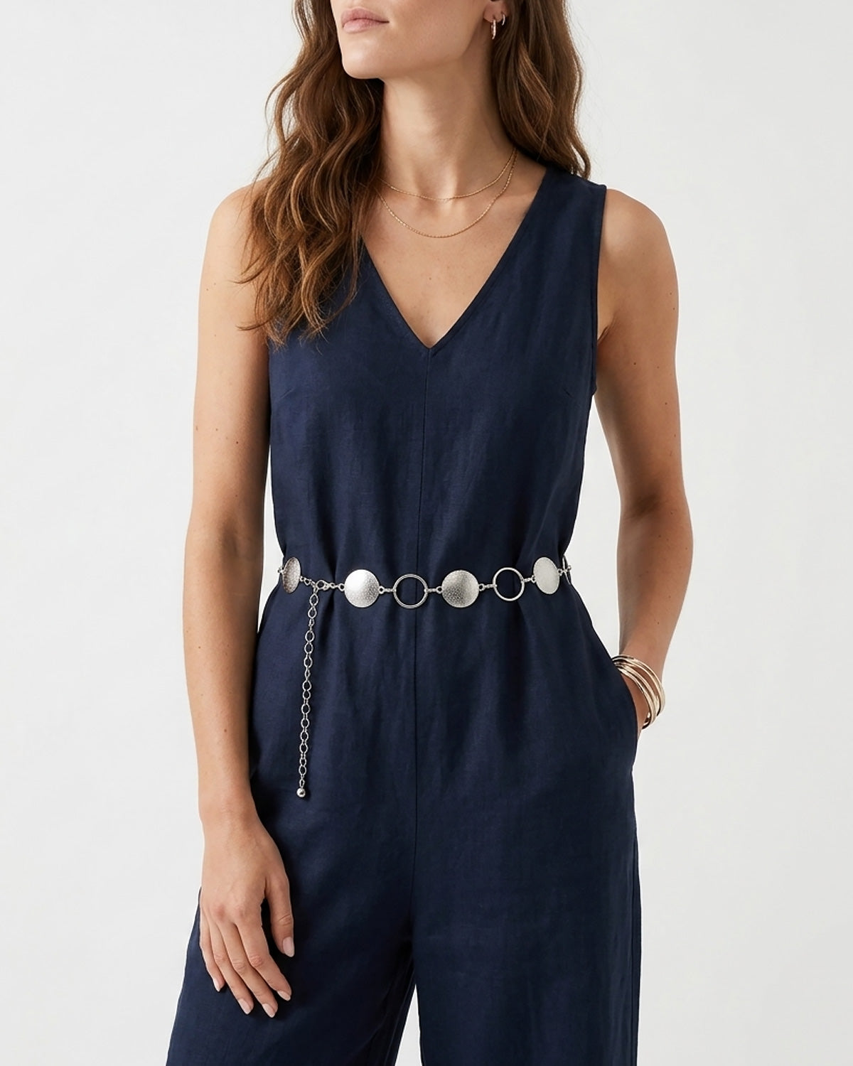 A woman wearing a navy blue sleeveless jumpsuit with a V-neckline and a decorative silver chain belt with rings and discs | BUCKLE Collective