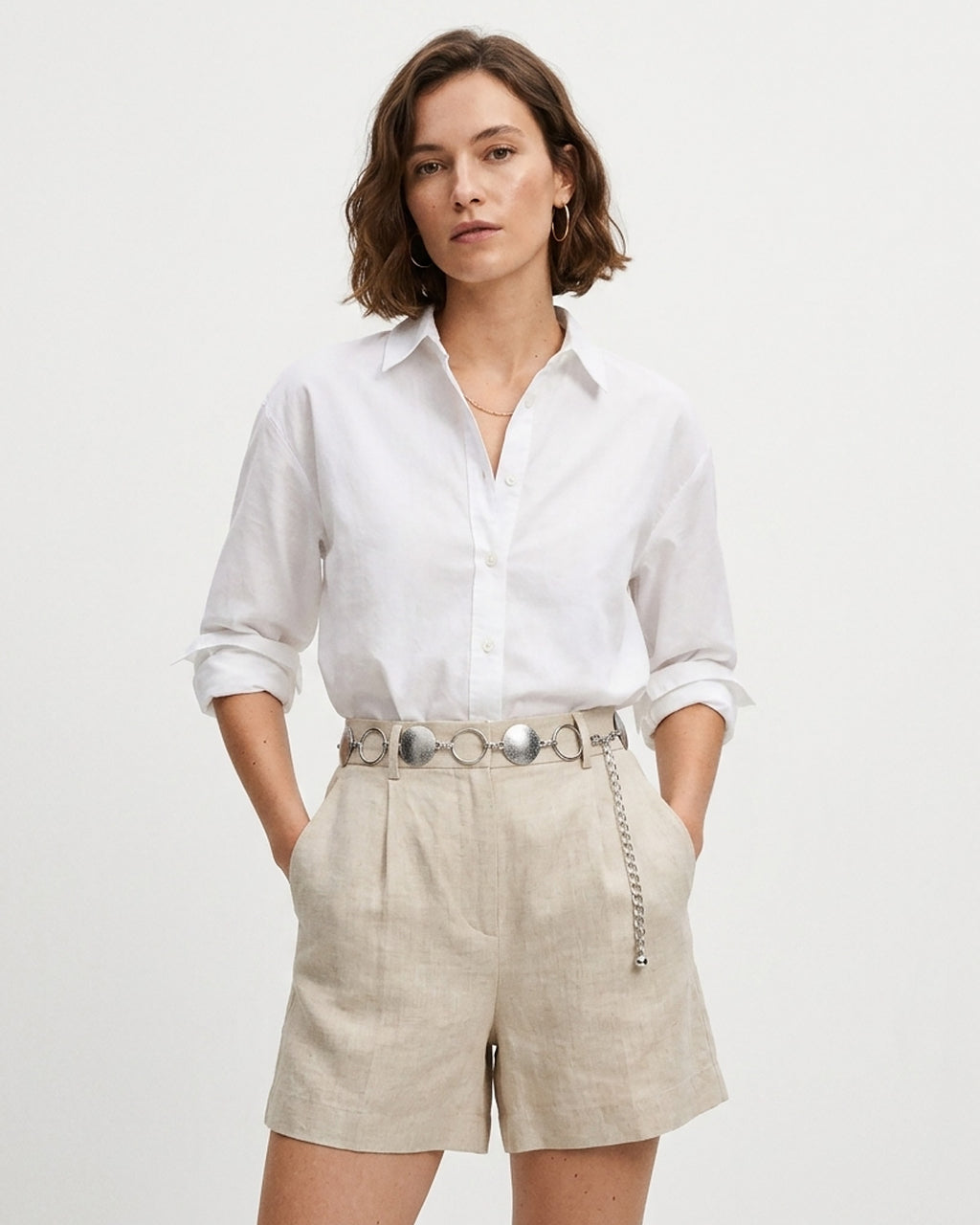A woman modeling a white long-sleeve button-up shirt and light beige linen shorts with a silver disc-ring chain belt | BUCKLE Collective