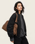 A woman wearing a black bomber jacket and gray sweater, carrying a brown studded shoulder bag 