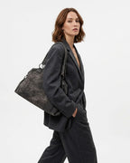 A woman wearing a dark charcoal gray blazer and matching trousers carries a large, distressed dark gray shoulder bag with metal studs against a plain white studio background.