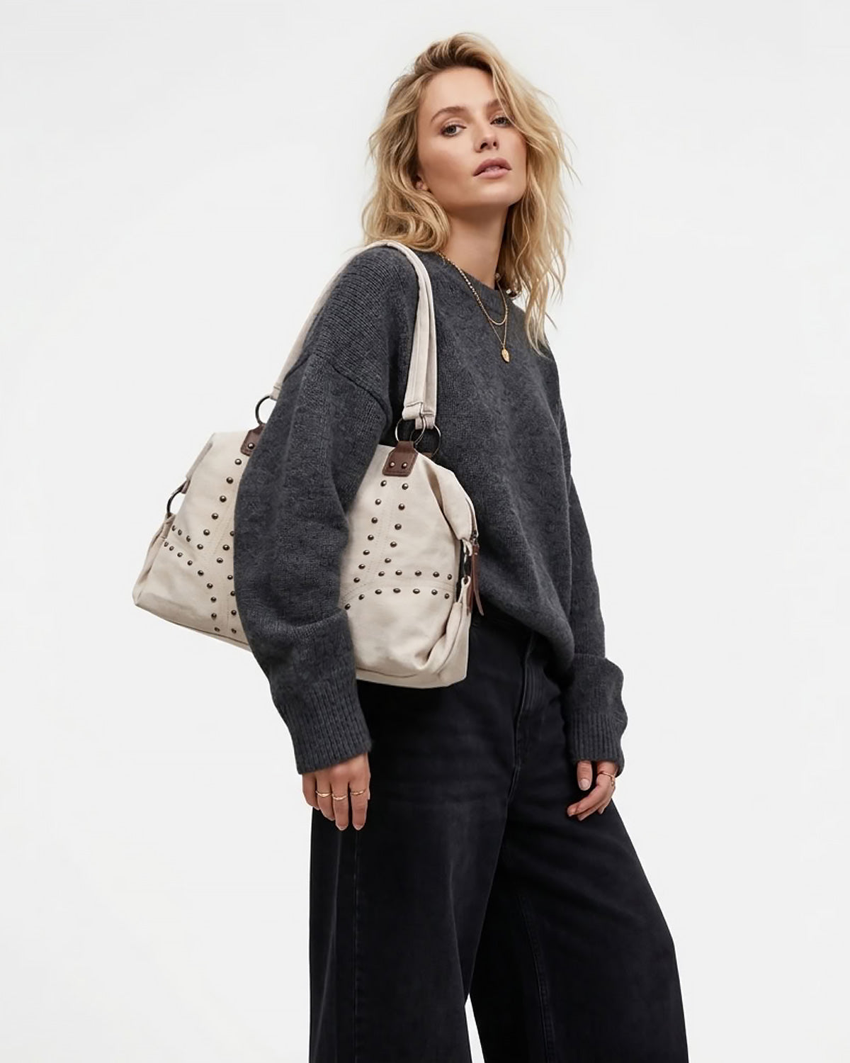 A woman wearing a dark gray knit sweater and black wide-leg pants carries a cream-colored studded shoulder bag with brown leather details