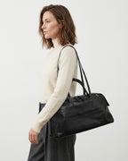 A woman in a cream turtleneck sweater and dark gray trousers carries a large black leather duffel bag over her shoulder | BUCKLE Collective