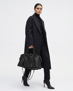 A model wearing a long black coat, black turtleneck, and black trousers, holding a black leather duffel bag | BUCKLE Collective