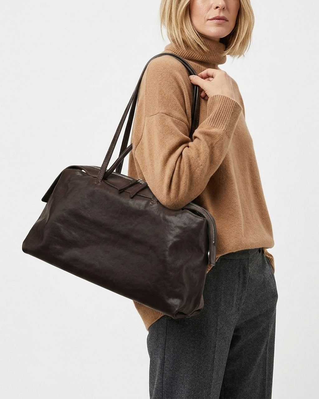 A woman wearing a camel turtleneck sweater and dark gray trousers carries a Large Coffee-brown Leather Pillow Duffle Handbag with Shoulder Straps | BUCKLE Collective