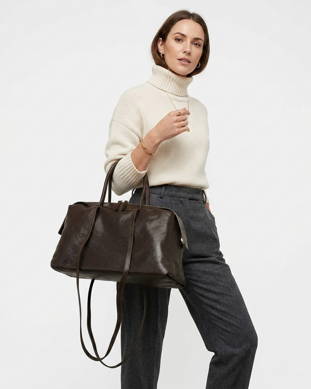 A woman wearing a cream turtleneck sweater and dark gray trousers carries a Large Coffee-brown Leather Pillow Duffle Handbag with Shoulder Straps | BUCKLE Collective