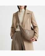 A model wearing a beige wool coat, cream turtleneck, and taupe trousers, carrying a large, crescent-shaped taupe lambskin leather crossbody | BUCKLE Collective