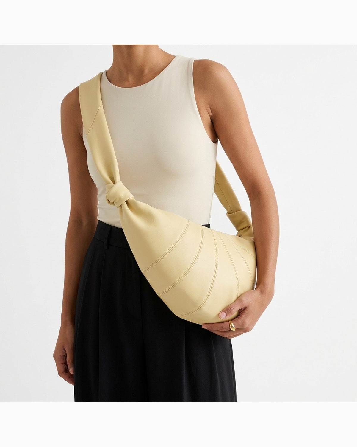 A model wearing a cream ribbed tank top and black pleated skirt carries a pale yellow knotted large lambskin crescent-shaped crossbody bag with a wide strap | BUCKLE Collective