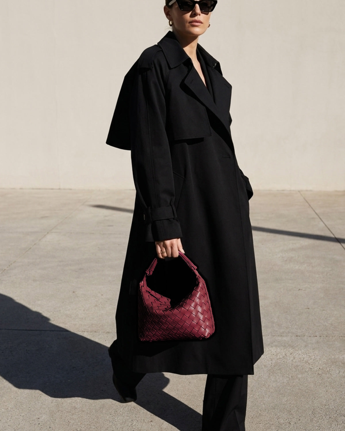 A stylish woman in a fashionable black coat carries a trendy Vegan Leather Woven HOBO Handbag with an adjustable strap in Wine Red color