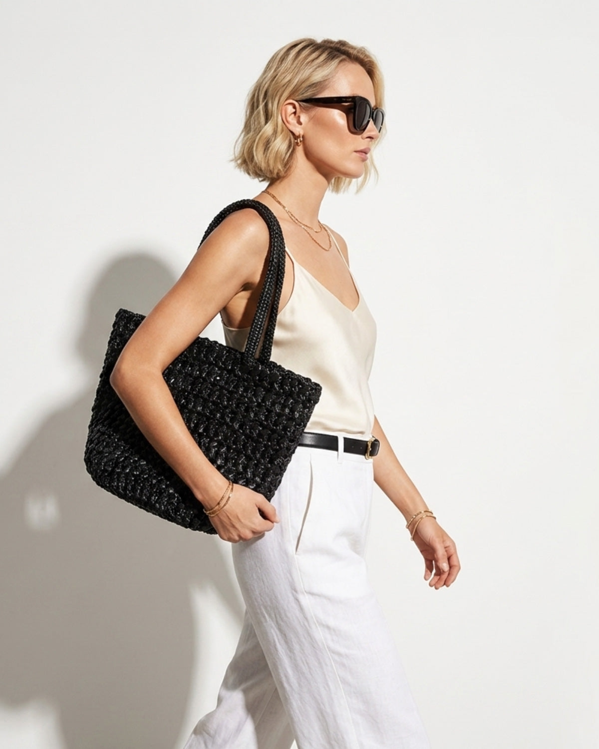 An elegant blonde woman in a sunglasses and stylish white-toned summer outfit with black belt, carries a Natural Black Woven Raffia Tote Bag with Removable Pouch