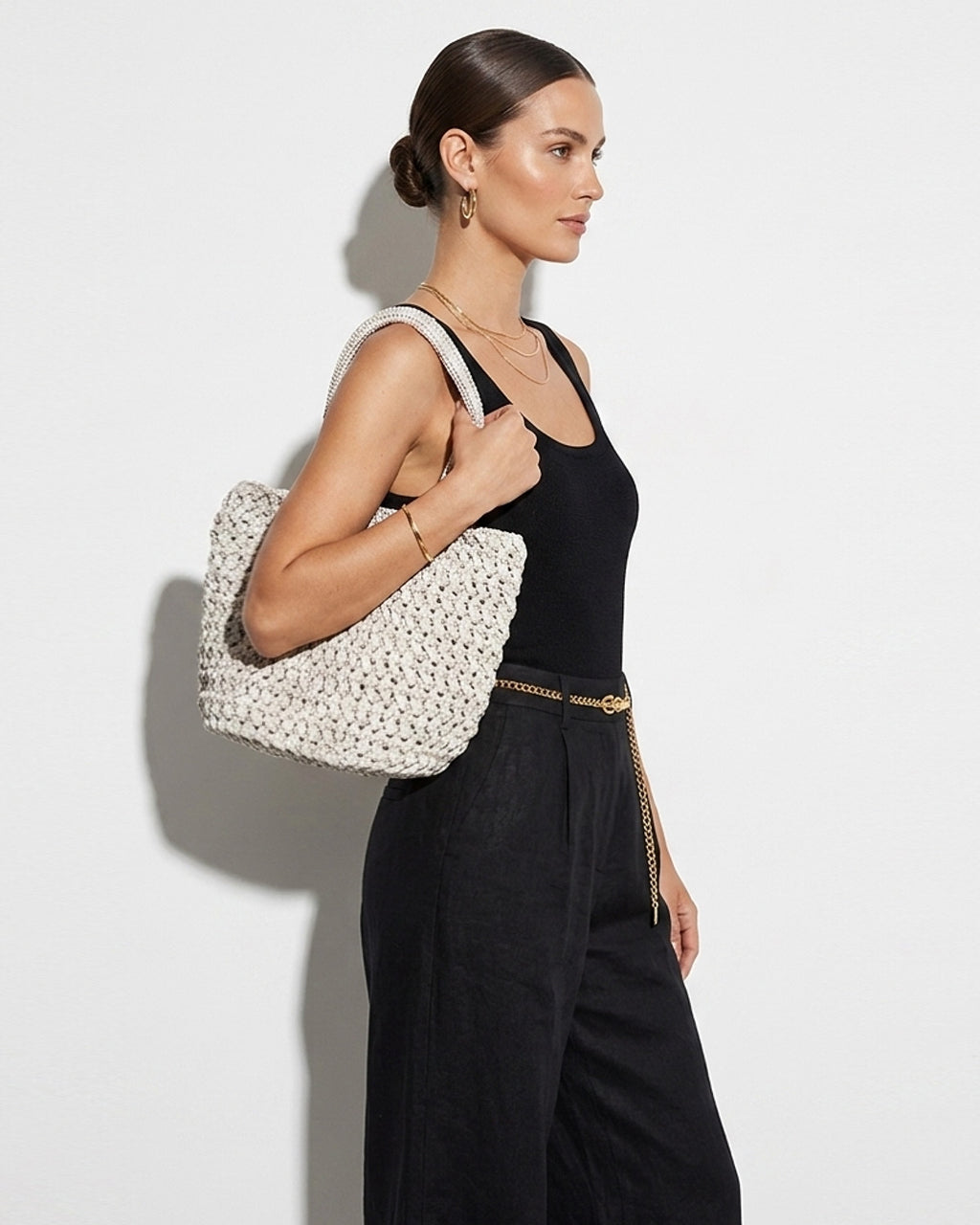 A stylish woman in a summer outfit, a black top and linen pants with a chain belt, carries a Natural Off-White Woven Raffia Tote Bag with Removable Pouch