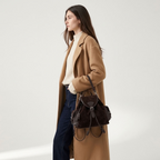 A woman wearing a camel coat, cream turtleneck, and dark blue denim holds an Espresso-Brown suede backpack | BUCKLE Collective