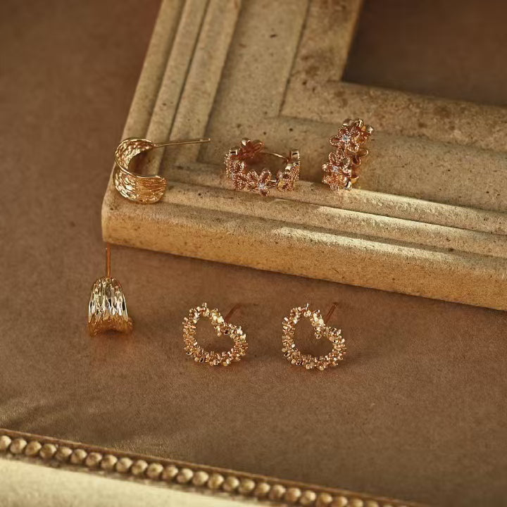 Gold-plated floral huggie hoop earrings with openwork four-petal flowers and sparkling cubic zirconia centres, displayed alongside other earrings on a vintage-style tray.