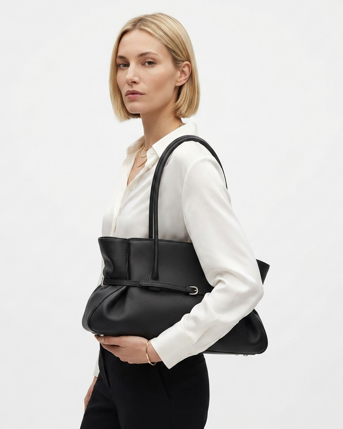 A blonde woman wearing a white blouse and black trousers carries a large black leather tote bag over her shoulder | BUCKLE Collective