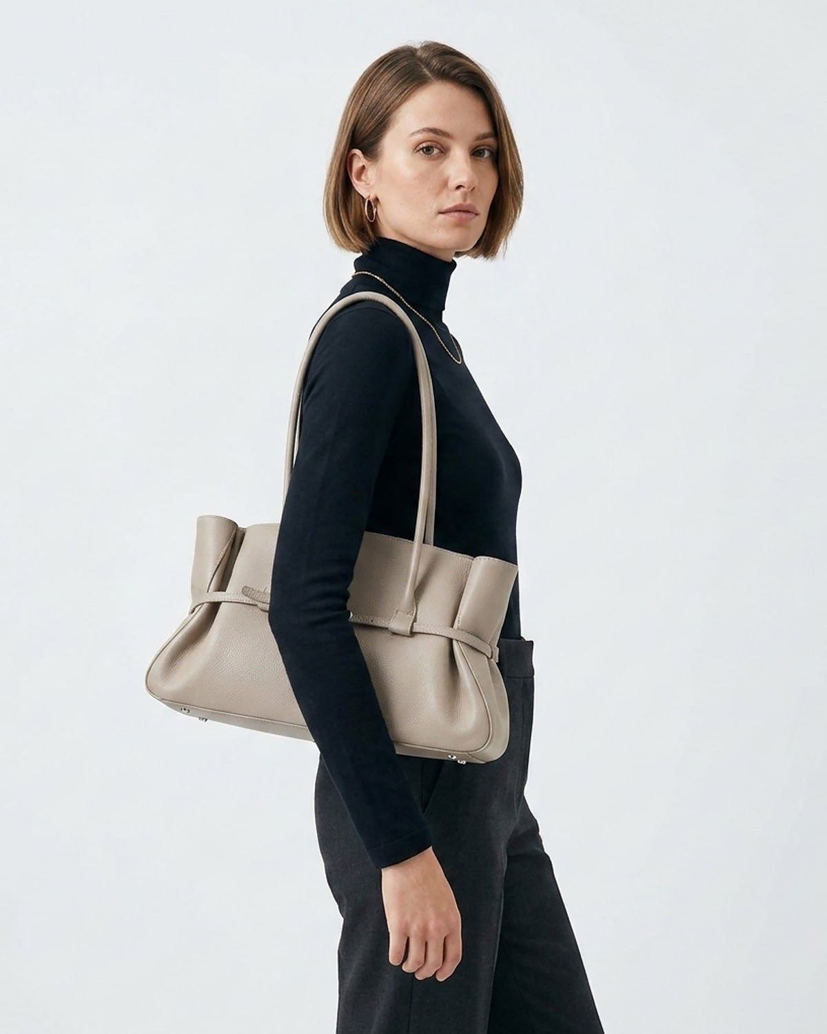 A woman in a navy blue turtleneck sweater and dark trousers carries a large taupe leather tote bag with double shoulder straps and a buckle strap detail | BUCKLE Collective